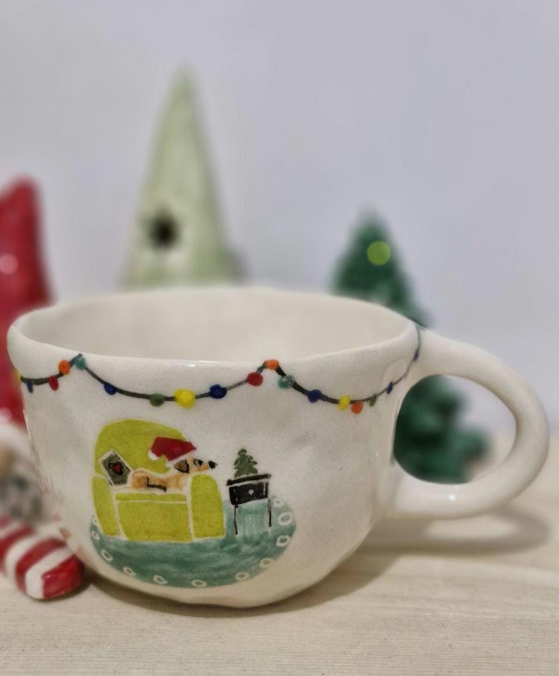 May include: White ceramic mug with a hand-painted design of a dog wearing a Santa hat sitting in a green armchair with a small Christmas tree next to it. The mug has a string of colorful lights around the rim.