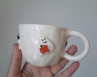 May include: A handmade white ceramic mug with a rounded handle. The mug features a smiling ghost and a red pumpkin decoration. The mug is held in a hand against a neutral background. The mug is a unique piece of pottery.