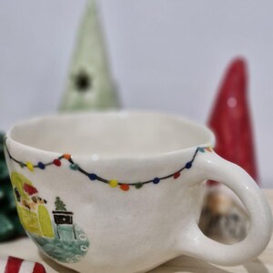 May include: White ceramic mug with a Christmas scene painted on it. The scene includes a small house with a Christmas tree, a snowman, and a string of lights. The mug has a handle and is decorated with a white glaze.