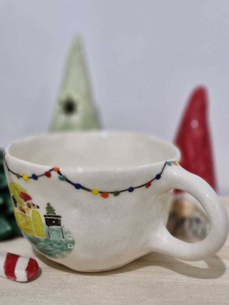 May include: White ceramic mug with a Christmas scene painted on it. The scene includes a small house with a Christmas tree, a snowman, and a string of lights. The mug has a handle and is decorated with a white glaze.