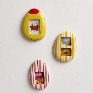 Può includere: Tre ornamenti da parete in ceramica con finiture lucide. Uno è giallo con una parte superiore rossa e un ritaglio rettangolare che mostra peperoncini rossi. Un altro è a righe gialle e bianche con due ritagli. Il terzo è a righe rosa e bianche.