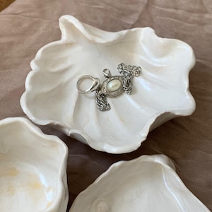 May include: White ceramic seashell-shaped trinket dishes. One dish holds a silver ring and a silver necklace with a pearl pendant. The dishes have a textured surface and are displayed on a neutral-colored background.
