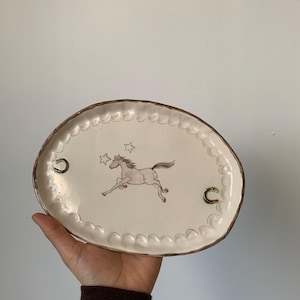 Puede incluir: Plato ovalado de cerámica con superficie crema y borde marrón. El plato presenta una ilustración de un caballo corriendo, dos estrellas y dos diseños de herraduras. El borde tiene un diseño festoneado.