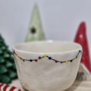 May include: White ceramic bowl with a string of colorful lights painted around the rim. The bowl is slightly textured and has a rounded shape.