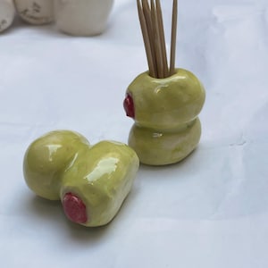Puede incluir: Porta palillos de cerámica con forma de aceituna, verde con detalles rojos, que contiene palillos de madera. También se ven dos aceitunas de cerámica. El diseño es temático de comida y está hecho a mano.