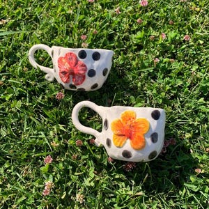 Può includere: Due tazze in ceramica bianca con pois neri e un fiore tridimensionale applicato sulla parte anteriore di ciascuna. Una tazza presenta un fiore rosso, mentre l'altra presenta un fiore arancione. Entrambe le tazze hanno un manico curvo sul lato.