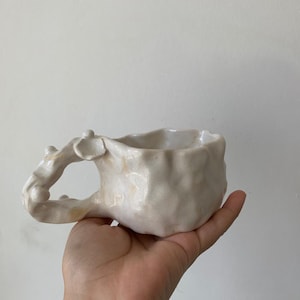 Könnte beinhalten: Eine handgefertigte, cremefarbene Keramiktasse mit organischem, strukturiertem Design. Die Tasse hat einen einzigartig geformten Henkel und einen leicht unregelmäßigen Rand. Die Tasse wird in einer Hand gehalten, um ihre Größe und Form zu zeigen. Das Innere der Tasse ist weiß.