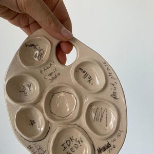 Pode incluir: Uma paleta de pintura de cerâmica creme com sete poços e uma alça. A paleta tem texto e desenhos manuscritos em preto, incluindo "PAKLAY Granic Studio", "Sketch First", "IDK RED?" e "Yellow".