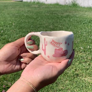 May include: A cream-colored ceramic mug with an irregular, hand-formed shape. The exterior features pink painted illustrations including a cactus, a cowboy hat, small stars, and the word LUCKY written in a stylized font above the hat design.