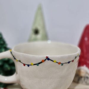May include: White ceramic mug with a string of colorful lights painted around the rim. The mug is sitting on a wooden surface with a candy cane and other Christmas decorations.