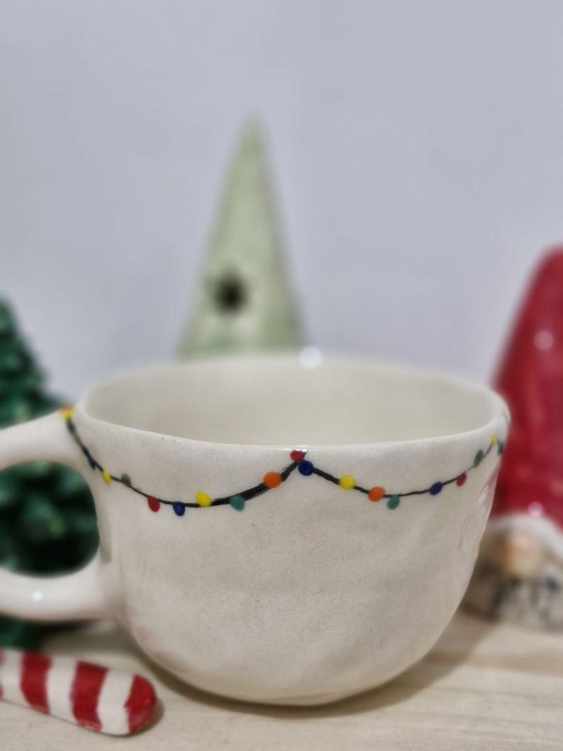 May include: White ceramic mug with a string of colorful lights painted around the rim. The mug is sitting on a wooden surface with a candy cane and other Christmas decorations.