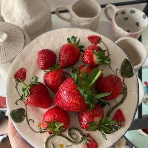Puede incluir: Un plato de fresas frescas y maduras con tallos y hojas verdes. El plato es blanco con un borde azul y un diseño de vid pintado. Otras piezas de cerámica están en el fondo, incluyendo tazas y un jarrón.