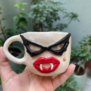 Peut inclure: Une tasse en céramique à base crème présentant un visage sculpté. La tasse arbore des lunettes œil-de-chat noires, des lèvres rouge vif avec deux crocs blancs dépassant vers le bas et un petit grain de beauté beige sur la joue droite.
