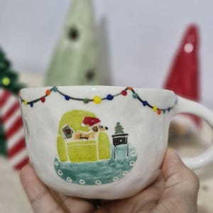 May include: White ceramic mug with a hand-painted design of a dog sitting in a chair with a Christmas tree and string of lights. The mug has a rounded shape and a handle.