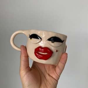 Op de afbeelding: Een beige keramische mok met een gezichtsontwerp. De mok heeft zwarte wimpers, zwarte wenkbrauwen en rode lippen. Hij heeft een oor en een klein gouden stipje aan de zijkant.