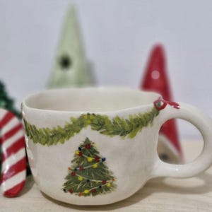 May include: White ceramic mug with a green garland and a green Christmas tree with red and yellow lights painted on it.