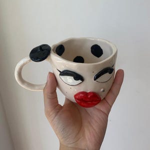 Op de afbeelding: Een crèmekleurige keramische mok met een handgeschilderd gezicht met dikke zwarte wenkbrauwen, lange wimpers en felrode lippen. Het handvat is versierd met een kleine zwarte baret en de binnenkant van de mok is voorzien van zwarte stippen.