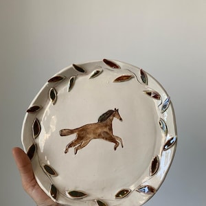 May include: A white ceramic plate with a brown horse illustration in the center. The plate is encircled by a decorative border of brown and green leaf-shaped accents. The plate is held by a person.