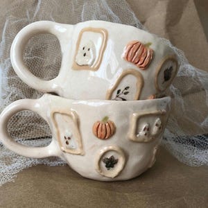 Peut inclure: Deux tasses en céramique empilées, de couleur crème. Chaque tasse présente des motifs en relief de citrouilles, de fantômes et de feuilles dans des cadres. Les tasses ont des anses arrondies, idéales pour l'automne ou Halloween.