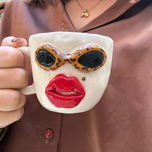 Può includere: Tazza in ceramica bianca con un viso dipinto a mano, con labbra rosse, occhi neri e occhiali da sole con stampa leopardata.