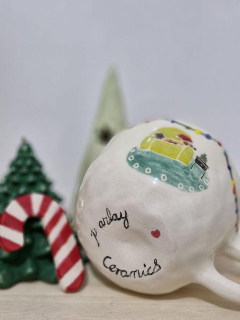 May include: White ceramic mug with a green, yellow, and red Christmas scene painted on the side. The mug has the words "p' arlay ceramics" written on the bottom.