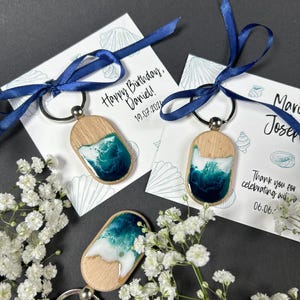 Könnte beinhalten: Holz-Schlüsselanhänger mit Kunstharz-Design im Ozean-Stil, in Blau-, Weiß- und Beigetönen. Jeder Schlüsselanhänger ist mit einem marineblauen Band an einer Karte befestigt. Die Karten haben Text.