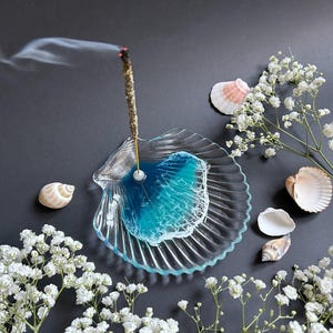 Op de afbeelding: Een heldere, schelpvormige wierookhouder met een blauw en wit harsontwerp, met een brandende wierookstok. De houder is omgeven door witte bloemen en schelpen op een donkere achtergrond.