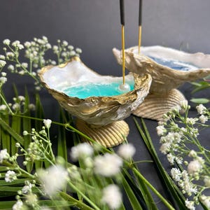Op de afbeelding: Twee witte schelpvormige wierookhouders met gouden accenten, gevuld met blauwe vloeistof en een parel. De houders staan op een zwarte ondergrond met groene bladeren en witte bloemen.