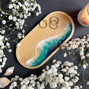 Op de afbeelding: Een houten ringenschotel met een hars-oceaanwave-ontwerp in tinten blauw, groen en wit. De schotel is ovaalvormig en heeft een natuurlijke houten afwerking. Drie zilveren ringen liggen op de schotel.