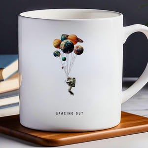 May include: White ceramic mug with a graphic of an astronaut holding onto a bunch of planets as if they were balloons. The text "SPACING OUT" is printed on the mug.