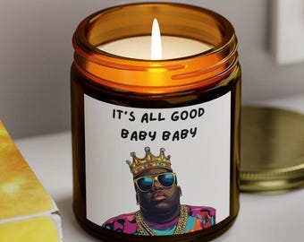 May include: A brown glass candle with a white label. The label features a cartoon image of a man wearing a gold crown, sunglasses, and a gold chain. The text on the label reads "It's all good baby baby".