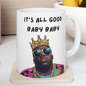 Peut inclure: Une tasse en céramique blanche avec une illustration en noir et blanc d'un homme portant une couronne dorée, des lunettes de soleil et des chaînes dorées. Le texte "IT'S ALL GOOD BABY BABY" est imprimé en noir sur la tasse.