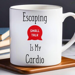 以下が含まれることがあります： 白いセラミック製のマグカップに、黒い文字で「Escaping Small Talk Is My Cardio」と書かれています。黒い文字で「Small Talk」と書かれた赤い吹き出しが、「Is My Cardio」という文字の上にあります。