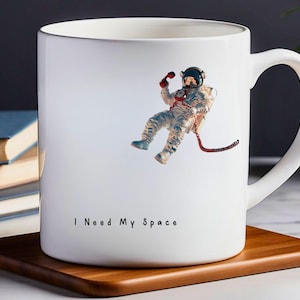 May include: White ceramic mug featuring a black and white image of an astronaut floating in space. The mug has the text "I Need My Space" printed on the bottom.