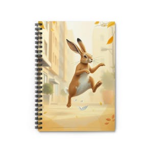 May include: A spiral-bound notebook featuring a cover illustration of a brown rabbit leaping through a city street. The rabbit has long ears, a white chest, and is surrounded by floating autumn leaves. The background shows blurred buildings.
