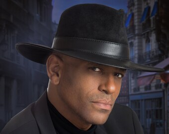 May include: A man wearing a black fedora hat and a black blazer. The hat has a black leather band. The man is looking directly at the camera. The background is a blurry image of a city street.
