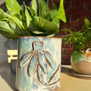 May include: A cylindrical ceramic planter with a raised floral design in shades of blue, green, and brown. The planter holds a lush green plant with variegated leaves. Another smaller planter with a fern is visible in the background.