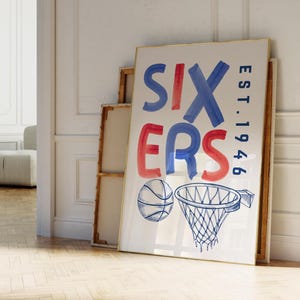 May include: A framed basketball-themed art print. The artwork features the word "SIXERS" in blue and red watercolor lettering, with "EST. 1946" in blue. A basketball and hoop are illustrated below. The frame has a gold border.