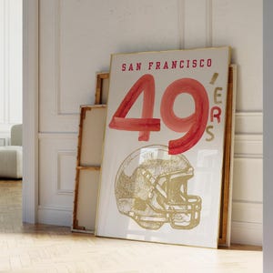 May include: A framed poster featuring the words "SAN FRANCISCO 49ERS" in red and gold. The number "49" is prominently displayed in red watercolor style, with a gold football helmet illustration below.