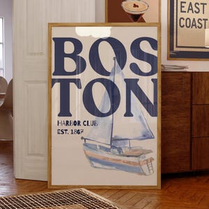 May include: A framed print with the word "BOSTON" in navy blue, above a watercolor sailboat illustration. The text "HARBOR CLUB EST. 1867" is below. The print is in a room with other framed art and wooden furniture.
