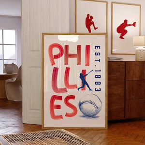 May include: A framed print with the word "PHILLIES" in red watercolor lettering, a blue baseball player silhouette, and a detailed baseball illustration. The print also includes "EST. 1883" in blue. Two additional framed prints with red baseball player silhouettes are visible.