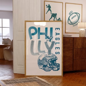 May include: A framed print with the words "PHILLY EAGLES" in teal blue, featuring a football helmet and an eagle illustration. Two smaller framed prints with football-themed silhouettes and a football graphic are also visible.