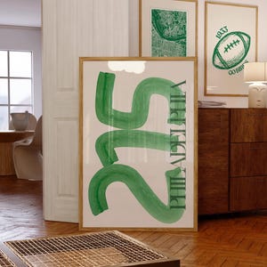 May include: A framed art print with the text "215 PHILADELPHIA" in green against a cream background. The artwork is in a wooden frame and is displayed in a room with other framed prints and wooden furniture.