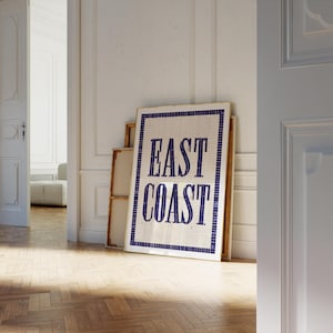 Könnte beinhalten: Ein weiß gerahmtes Schild mit blauen Mosaikfliesen, die "EAST COAST" in einer Vintage-Schriftart buchstabieren. Das Schild lehnt an einer weißen Wand mit einem Holzboden.