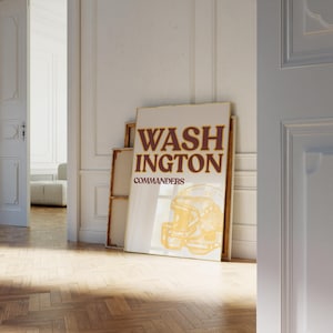 Pode incluir: Um poster emoldurado com o texto "WASHINGTON COMMANDERS" em letras bordô. Abaixo, um desenho de capacete de futebol americano dourado. O poster está encostado a uma parede em uma sala com piso de madeira.
