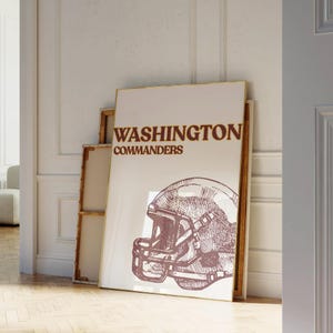 May include: A framed poster with the text "WASHINGTON COMMANDERS" in brown. Below the text is a detailed illustration of a football helmet. The poster is displayed against a white wall, with a wooden frame.