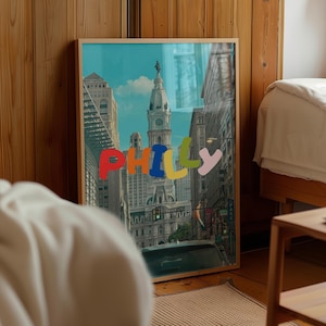 May include: A framed art print featuring a colorful cityscape of Philadelphia, with the word "PHILLY" in large, multi-colored letters across the image. The artwork captures a street view with buildings and a clock tower under a blue sky.