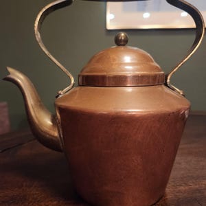 May include: A copper kettle with a wooden handle and a brass spout and handle. The kettle has a rounded body and a lid with a small knob. The kettle is sitting on a wooden surface.