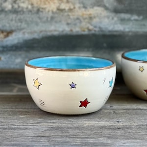 May include: Two off-white ceramic bowls with a light blue interior and star designs in various colors. The bowls are on a wooden surface. The text "Sold separately" is at the bottom of the image.