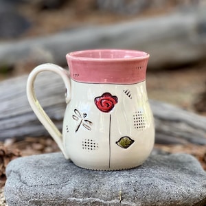 May include: A ceramic mug with a cream-colored body and a pink rim. The mug features a hand-painted red flower, a brown dragonfly, a green leaf, and small black grid patterns. It has a curved handle on the left side and a rounded base.